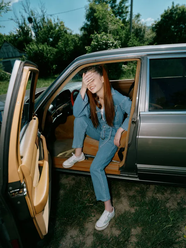 Young woman in denim outfit sitting in car, expressing joy and relaxation in a sunny outdoor setting, surrounded by green nature.