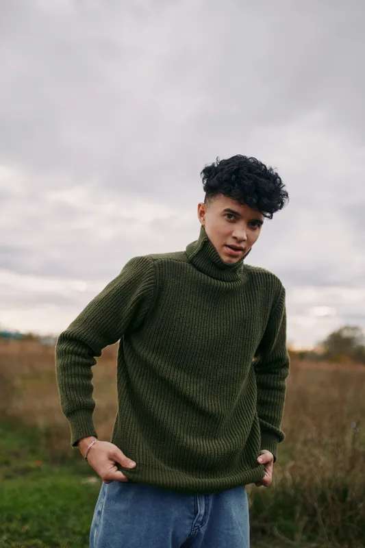 A young man stands in an open field wearing a chunky green turtleneck, relaxed pose, authentic everyday style, candid portrait under soft overcast light.