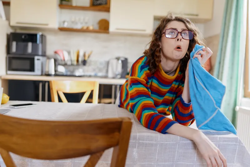 A woman in a colorful striped sweater stands at a kitchen table, wiping a surface with a blue cloth, glasses perched, surprised expression. Casual home life, everyday chores.