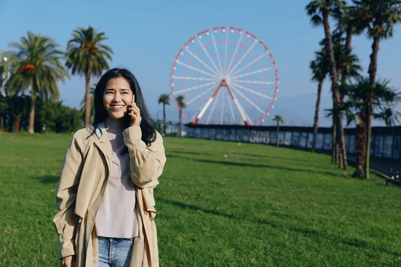 Woman in a beige coat smiles while talking on her phone in a sunny park, green grass and a distant ferris wheel creating a casual outdoor lifestyle scene