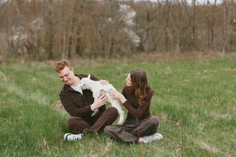 Cheerful couple outdoors, playing with a goat kid in a sunlit field, relaxed interaction, candid moment, natural colors, casual clothing, warm breeze, rustic backdrop and joyful smiles