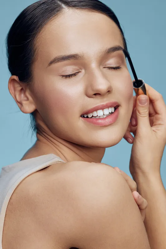 Young woman applying mascara with closed eyes, showcasing a natural beauty look against a soft blue background, highlighting flawless skin and subtle makeup