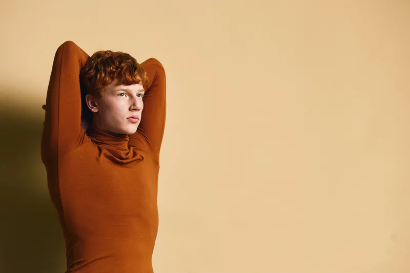Young man with red hair and curly hairstyle wearing brown turtleneck sweater posing with arms behind head against beige background with thoughtful expression and casual style.