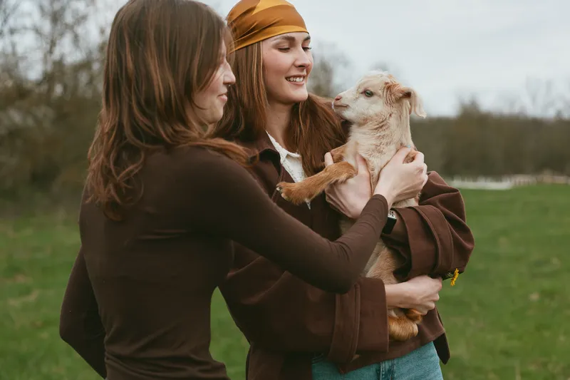 women outdoor goat kid friendship lifestyle, two women outdoors smiling softly with a young goat kid, pastoral meadow setting, casual cozy clothing, warm light, natural colors, affectionate