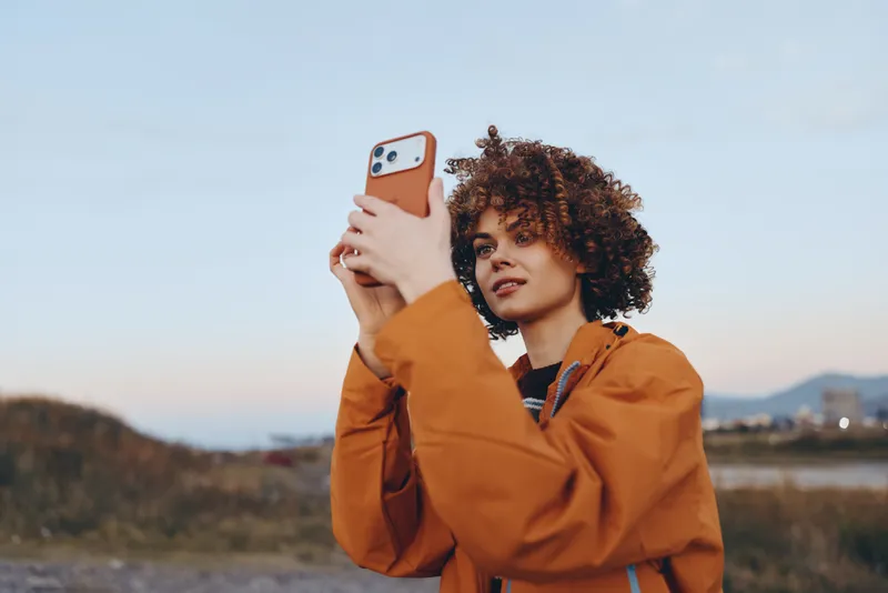 Woman lifestyle outdoors, curly hair and jacket, holding smartphone for selfie at sunset in a field, soft wind, golden warm light, calm cinematic portrait