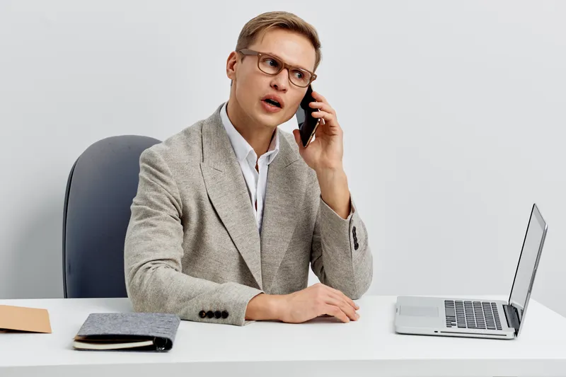 Businessman talking on the phone during work, sitting at desk with laptop, office environment, formal attire, serious facial expression, modern workplace, professional communication concept.