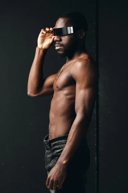 Creative conceptual portrait of a shirtless African man wearing futuristic black visor glasses against dark background. Visual style emphasizes modern fashion and technology.