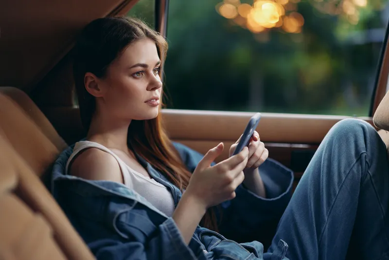 A thoughtful young woman with long hair in a denim jacket sitting in a car, engaging with her phone during sunset, highlighting modern lifestyle and technology.