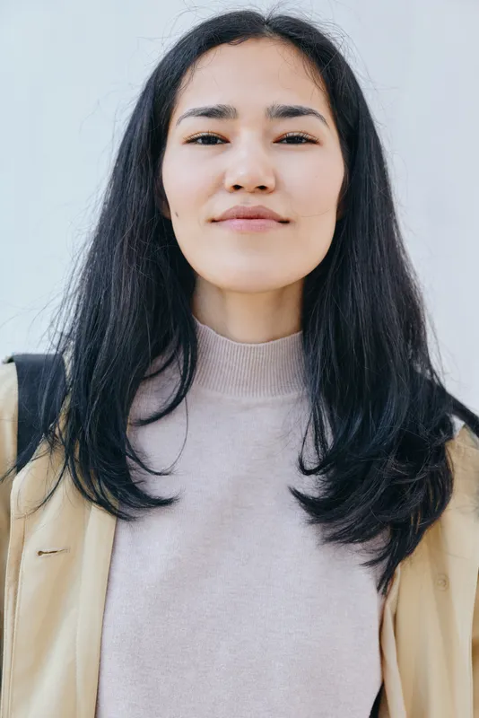 Casual woman standing against a pale backdrop with a confident expression, wearing a light sweater and beige jacket, natural lighting, everyday fashion, street style and candid context for editorial