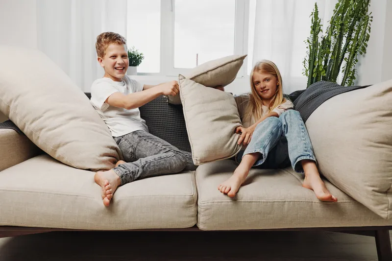 Two children having a pillow fight on a beige sofa in a bright living room with plants and natural light.