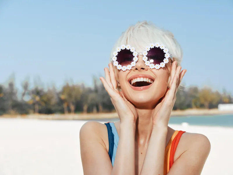 Playful sunlit beach scene featuring a smiling woman with quirky flower sunglasses, bright swimsuit straps, and a carefree mood, capturing youthful summer joy and confidence.