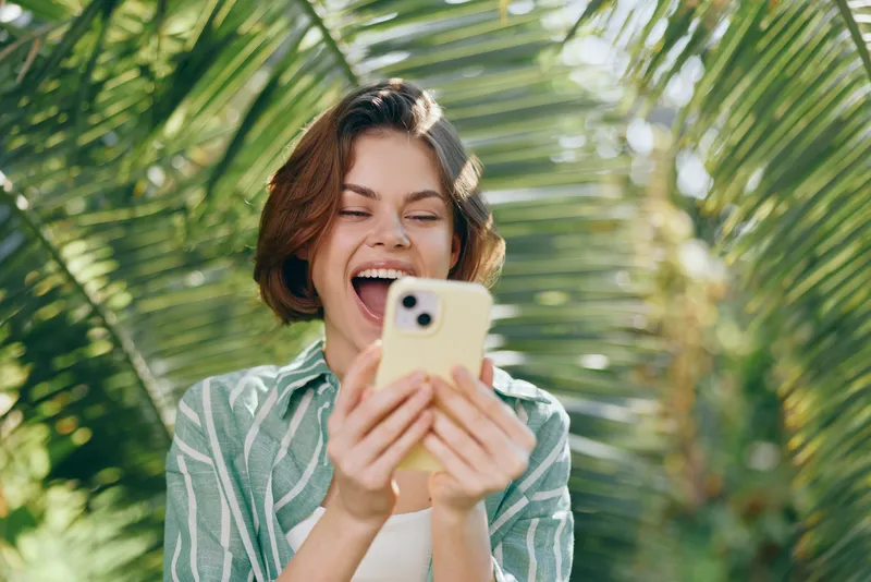 Young woman smiling brightly as she looks at her smartphone outdoors among tropical plants.