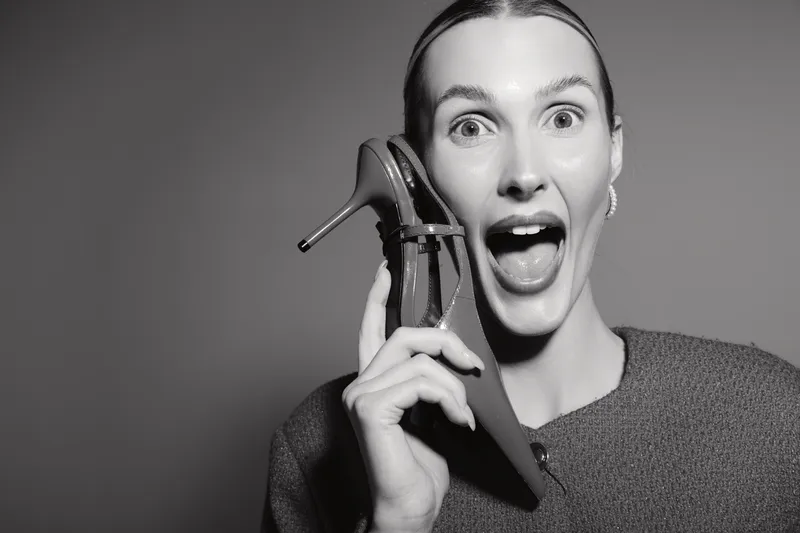 Smiling white woman with fair skin holds high heel shoe near face with open mouth. Studio portrait on grey background.