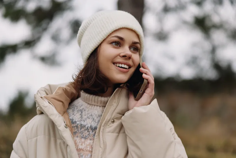 Woman outdoors in a warm jacket and beanie, talking on a smartphone and smiling, with a soft breezy landscape and distant trees.