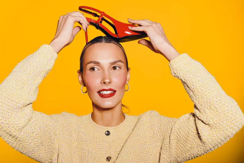 Young woman with red lipstick holds red high heel shoe above head on bright yellow background. Fashion concept with vibrant colors and playful mood in studio lighting.