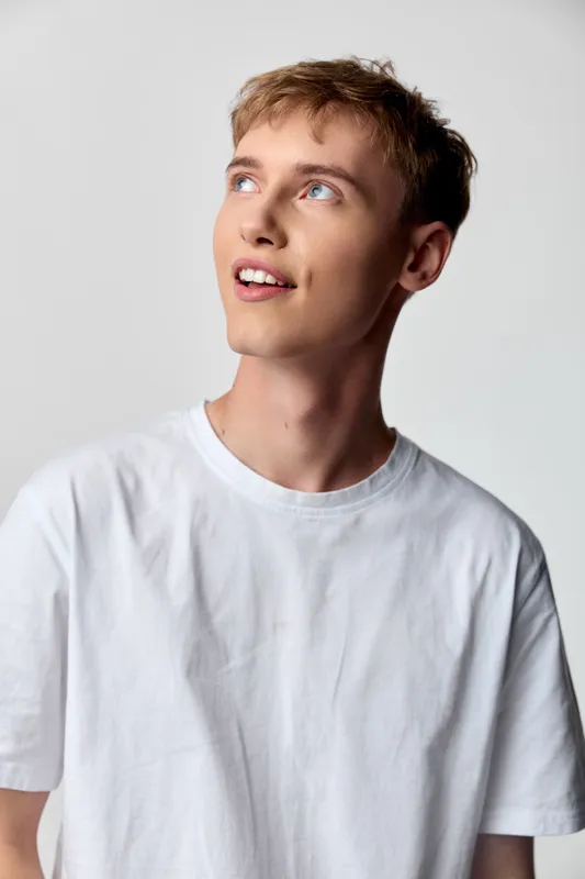Young man in a white tee gazing upward with a curious smile, casual and thoughtful, studio lighting highlights fashion and lifestyle details in a clean minimal setting