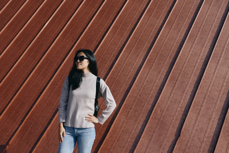 Woman wearing sunglasses, denim jeans and a sweater, with a backpack, standing confidently against a diagonal metal wall in an urban outdoor setting, showcasing casual street fashion and bold attitude