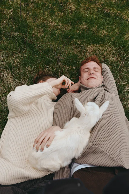 Couple lying on grass in cozy sweaters with a goat kid between them, sharing a calm outdoor moment, relaxed smiles, natural light, lifestyle portrait, intimate countryside vibe, soft breeze.