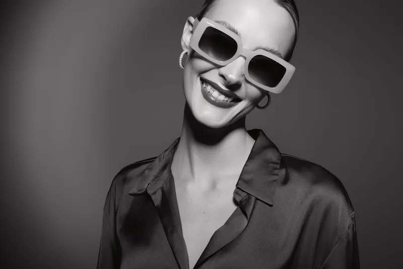 Smiling Caucasian woman with light skin wearing large sunglasses and silky blouse poses in studio with grey background. Concept of fashion, style, and confident female model.