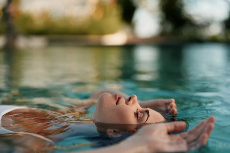 Relaxed woman floating on water surface in outdoor pool, eyes closed, peaceful expression, natural light, calm atmosphere, summer lifestyle, serene moment