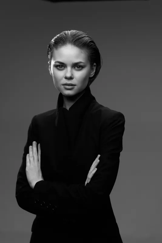 Black and white portrait of serious woman in dark suit with crossed arms against plain background.