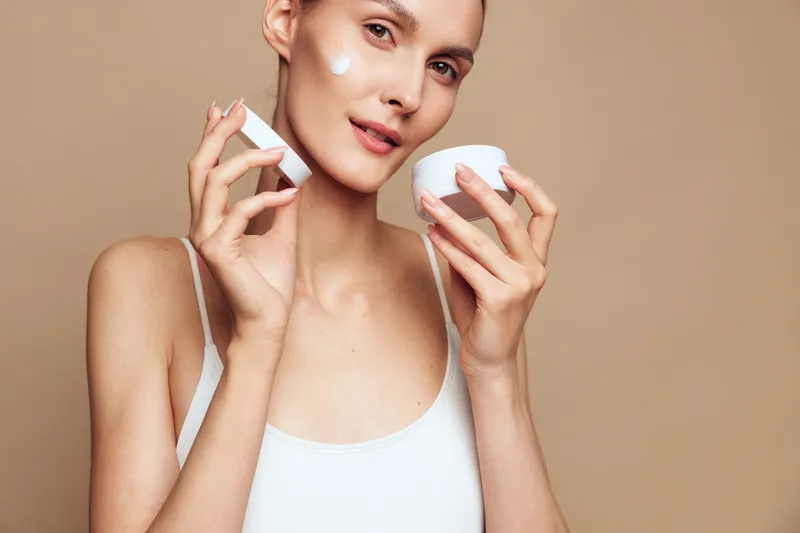 A woman applies moisturizer from a jar while holding a small bottle, smiling softly in a clean, neutral setting. Concept: skincare routine and self care.