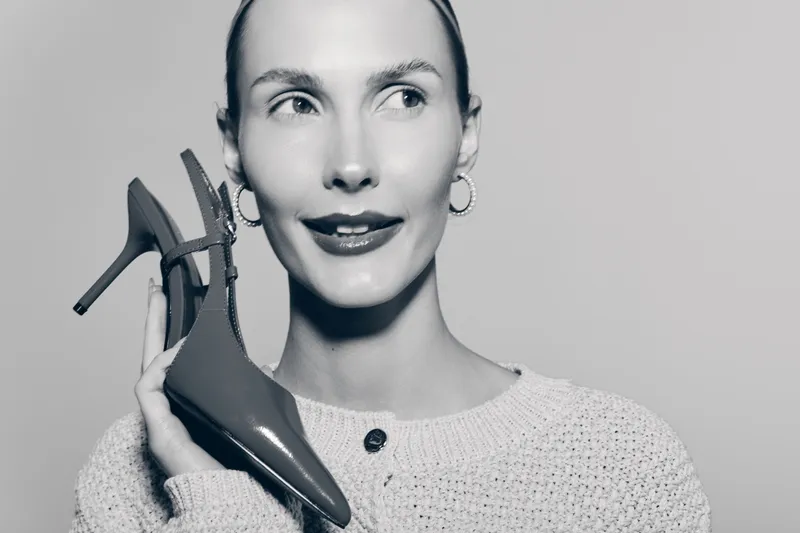 A smiling white woman with fair skin holds a high heel shoe near her face. Studio portrait with fashion concept and elegant style.