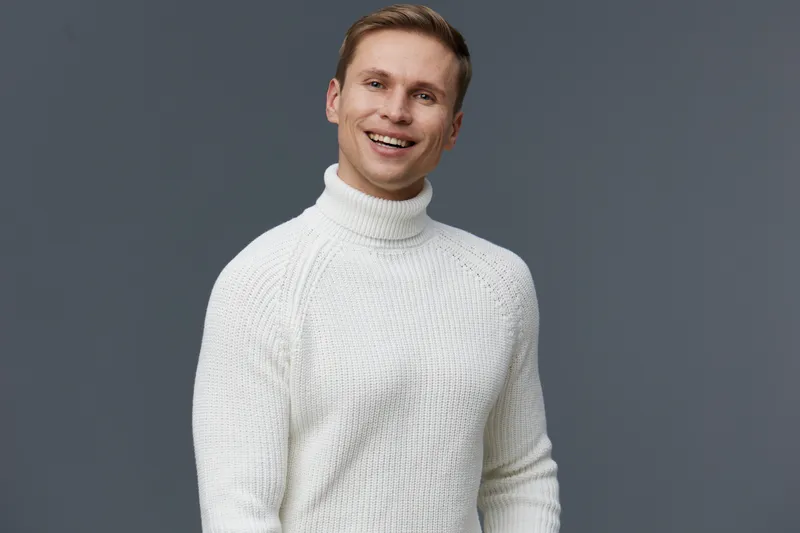Smiling man in white knitted turtleneck sweater looking at camera on plain gray background studio portrait. People lifestyle concept enjoying happy moment and warmth, male adult cheerful friendly fash