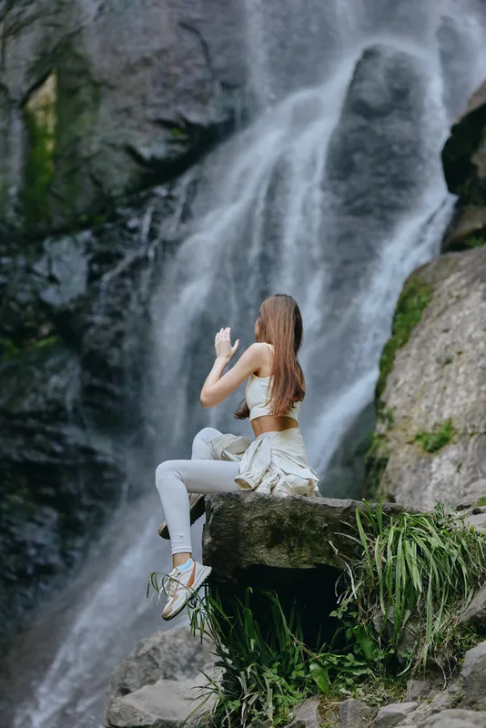 Woman meditating in white activewear sits peacefully on rock near waterfall surrounded by green nature, conveying relaxation and mindfulness in outdoor environment.