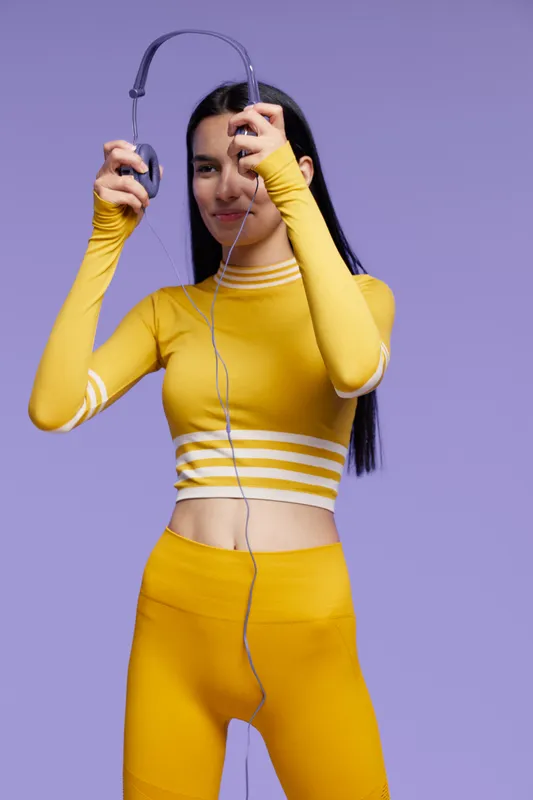 Woman sport fitness wearing yellow activewear, holding headphones with a joyful expression against a solid purple background, promoting health and energy.