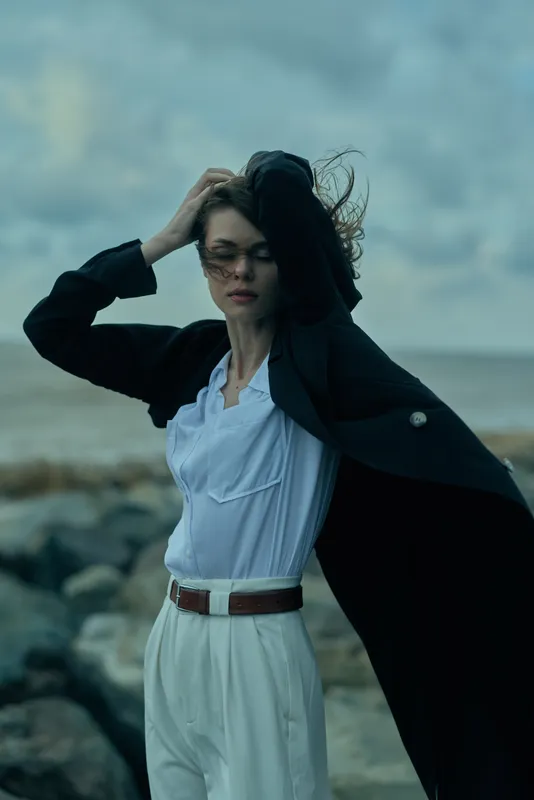 Fashion model by the sea, wind-swept coat and light blue shirt over white trousers, dramatic coastal look with bold, cinematic movement
