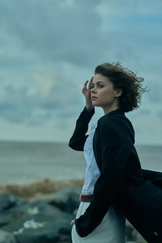 Windy coast scene, woman in blazer and white dress, gazing toward the horizon, dramatic clouds over the sea, rugged rocks