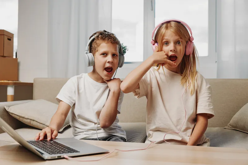 Two kids with headphones using laptop at home sitting on sofa, showing playful and surprised expressions. Concept of technology, fun, childhood, and entertainment.