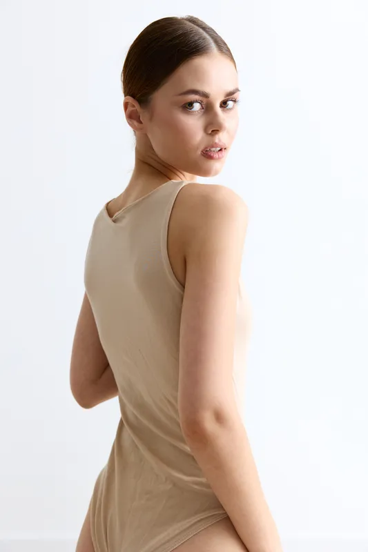 Young woman in beige sleeveless bodysuit looking back over shoulder against white background, natural light, minimalistic style, thoughtful expression, clean composition.