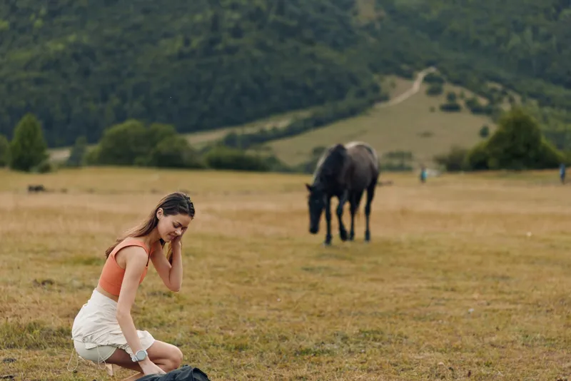 A cheerful young woman sits in a grassy field as a horse grazes nearby, enjoying a peaceful outdoor moment surrounded by open hills and calm countryside, creating a natural atmosphere.
