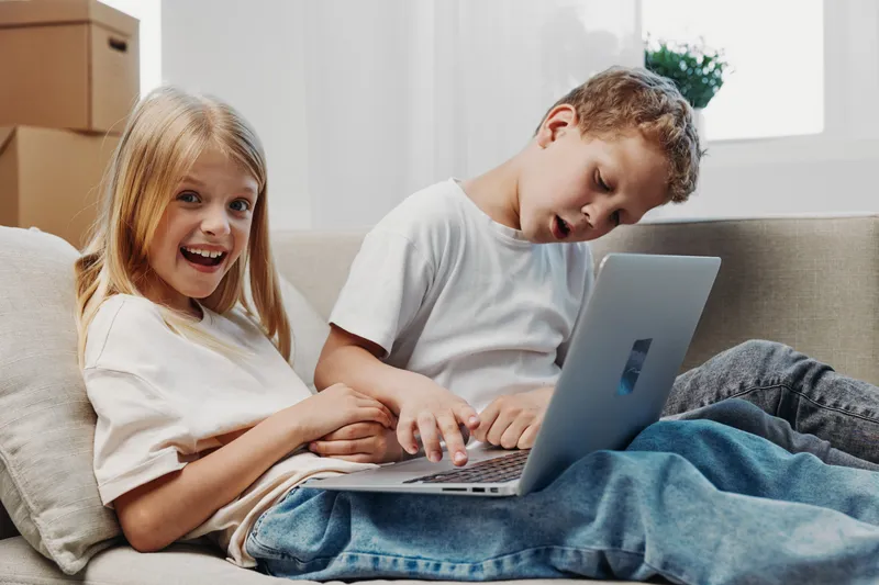 Two children sitting on a sofa using a laptop together, girl smiling excitedly while boy focuses on the screen. Concept of technology, learning, and childhood interaction.