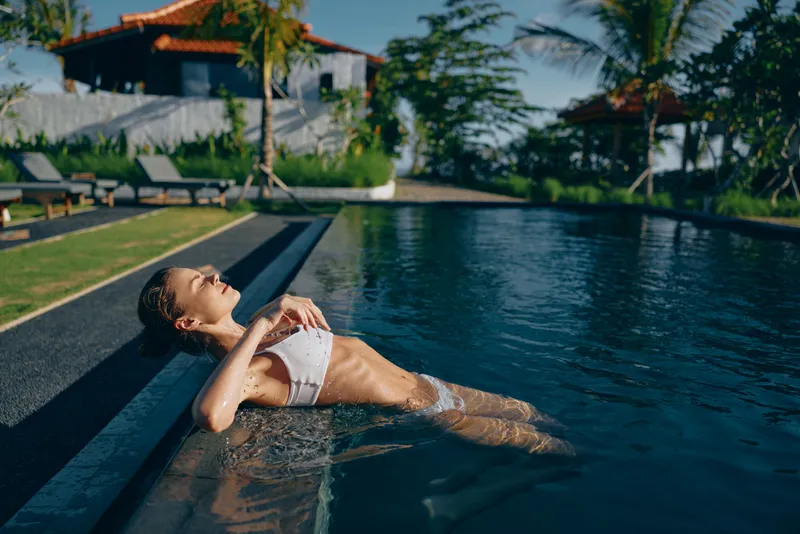 Young woman in white bikini relaxing in outdoor pool near tropical garden and lounge chairs. Lifestyle scene with sunny weather and calm water, concept of leisure and summer vacation.