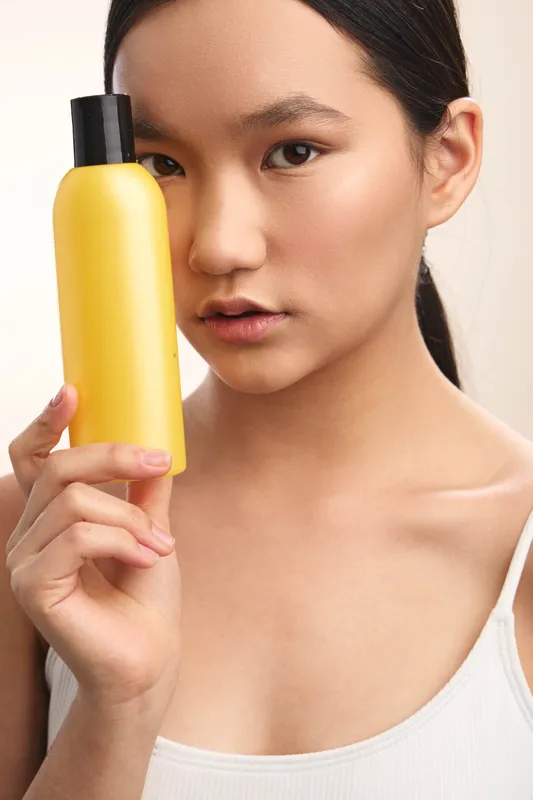 asian woman holds a yellow bottle close to her face in a clean studio setting, showcasing skincare product with a calm expression, soft lighting, and a minimal beauty vibe.