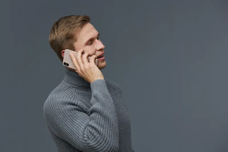Young caucasian man talking on smartphone wearing grey knitted sweater isolated on dark gray background side view communication concept digital nomad technology