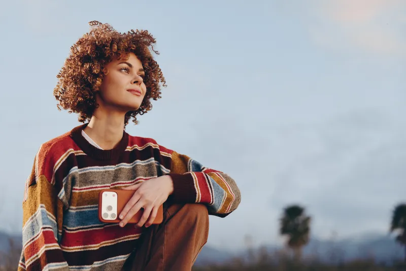 Woman with curly hair wears striped sweater outdoors holding smartphone near chest, lifestyle travel moment, sunset sky and distant mountains blurred, calm expression, casual style
