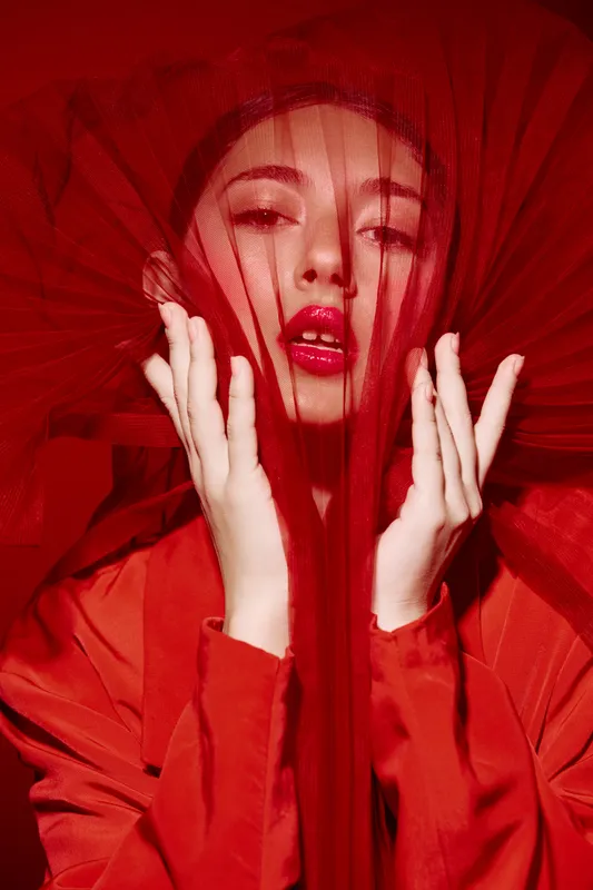 Fashion model with red lipstick and vibrant outfit gazes through sheer red fabric. Creative beauty concept with dramatic lighting highlighting glossy lips and delicate hands.