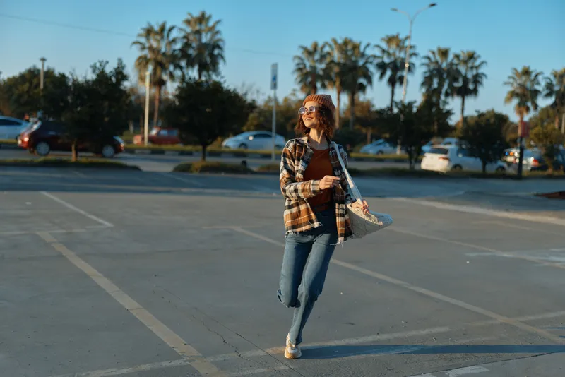 Woman walking coffee smile authenticity candid lifestyle urban: young woman walks across a sunlit parking lot with coffee and tote, golden hour glow, mindful living, emotional storytelling.