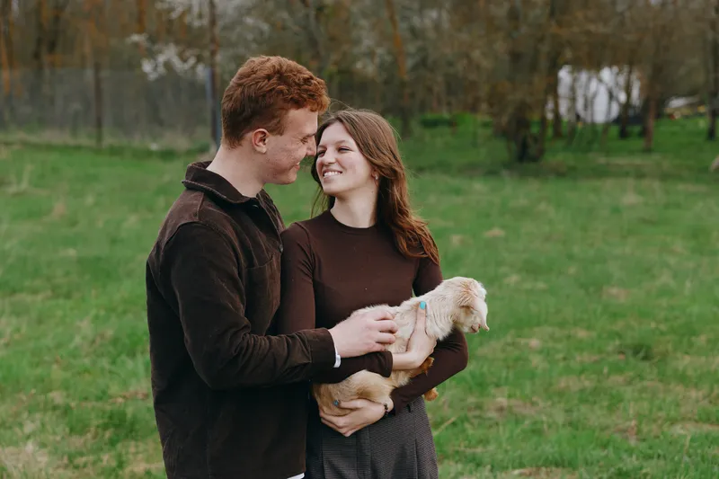 Young couple in casual clothes enjoys holding a baby goat together outdoors in a green field while sharing a tender moment and smiling at each other.