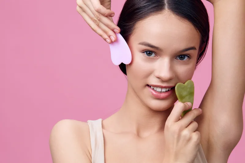 Beautiful young woman using facial tools for skincare, showcasing a healthy lifestyle with green and pink stones against a soft pink background