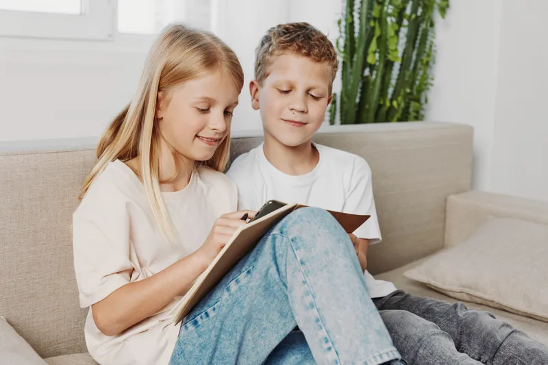 Happy children sitting on sofa, reading and writing in notebook together. Cozy home interior with natural light and plants in background.