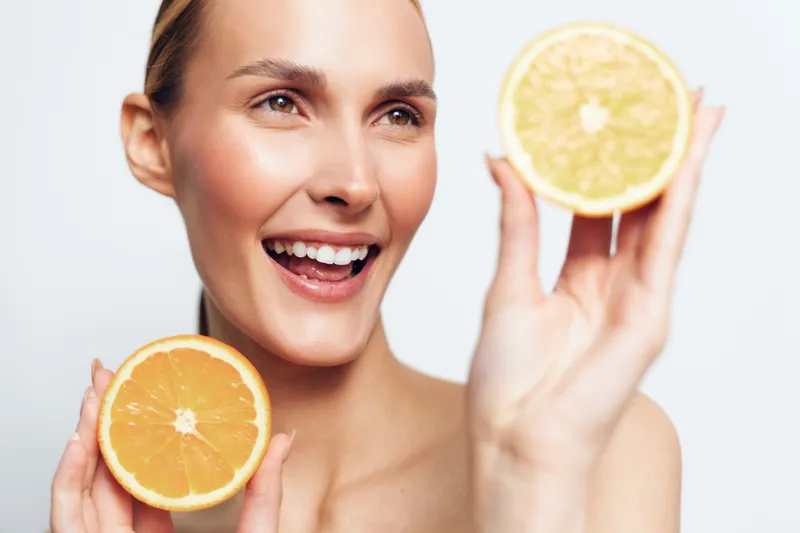 Happy woman holding two orange halves near face, smiling brightly, fresh fruit concept, natural skin, closeup portrait, white background, healthy lifestyle, vitamin C source