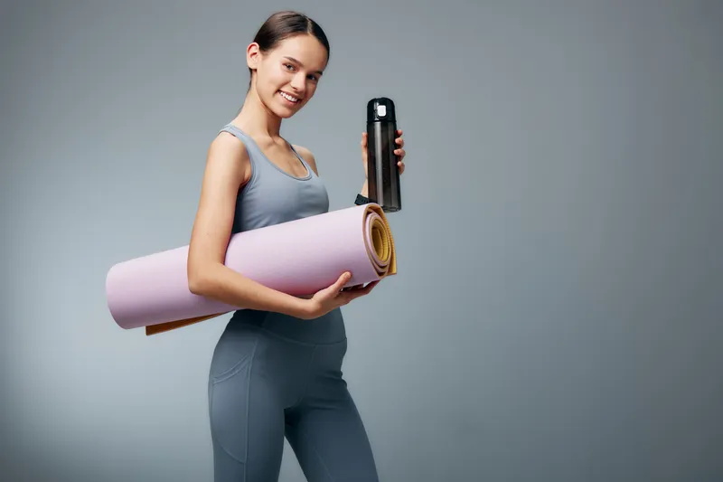 Fitness woman in athletic wear carries a yoga mat and a water bottle, ready for training. Modern studio backdrop with neutral tones highlights health, exercise and motivation. Her fit frame and