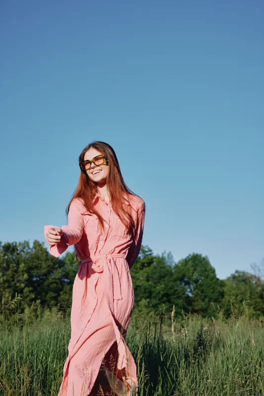 Young woman walking in a pink dress with sunglasses in a sunny green field, exuding joy and confidence against a bright blue sky