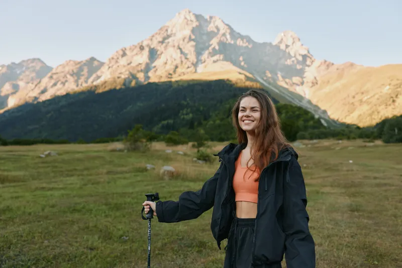 A cheerful woman outdoors wearing a jacket and athletic top stands with a trekking pole in a rugged landscape. She smiles warmly, radiating energy and adventure.