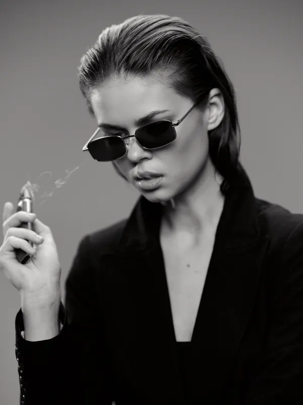 Black and white portrait of a woman wearing sunglasses and a blazer holding a cigarette, with a serious expression.